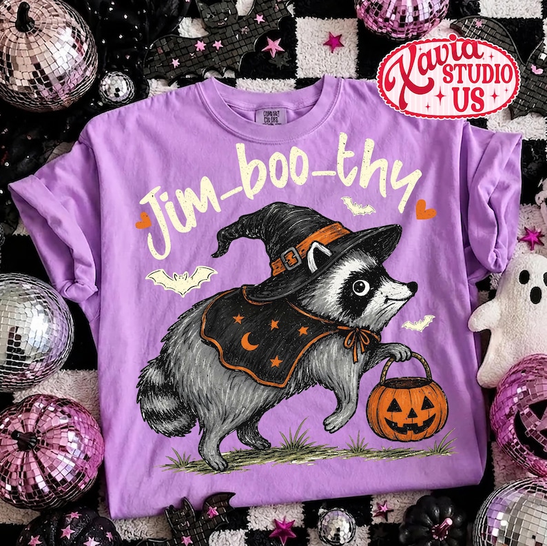May include: A light purple t-shirt featuring a graphic of a raccoon wearing a black witch hat and a cape with orange stars and a crescent moon. The raccoon holds an orange jack-o'-lantern bucket. The text Jim-boo-thy is printed in yellow.