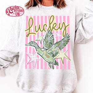 May include: A light gray sweatshirt featuring a graphic design with the word "Lucky" in green script. The design includes a green and white duck with star accents on a pink striped background.