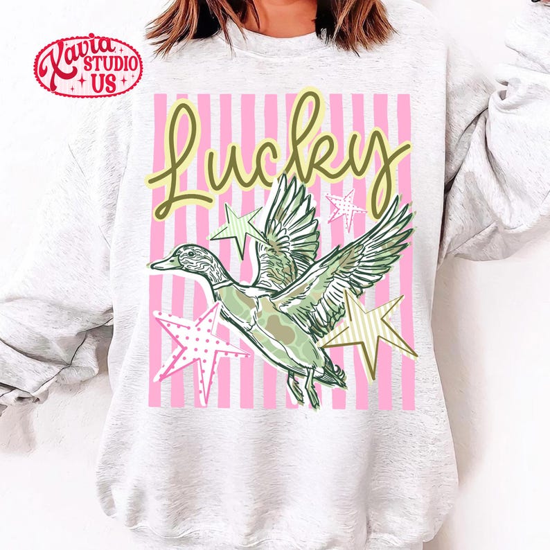 May include: A light gray sweatshirt featuring a graphic design with the word "Lucky" in green script. The design includes a green and white duck with star accents on a pink striped background.