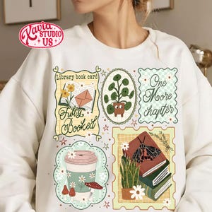 May include: White sweatshirt with multiple graphic designs. The designs include a coffee cup, a stack of books with a moth, a potted plant, and the phrases "Fully Booked" and "One More Chapter". The sweatshirt has a crew neck.
