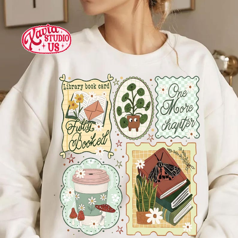 May include: White sweatshirt with multiple graphic designs. The designs include a coffee cup, a stack of books with a moth, a potted plant, and the phrases "Fully Booked" and "One More Chapter". The sweatshirt has a crew neck.