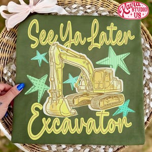 Pu&ograve; includere: T-shirt verde con grafica di escavatore giallo e la scritta "See Ya Later Excavator". Il design include stelle gialle e turchesi. La maglietta &egrave; esposta su una superficie tessuta.