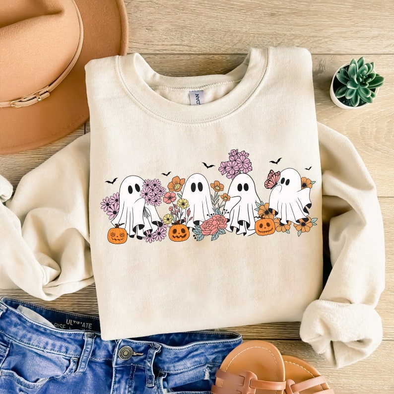 May include: A cream-colored sweatshirt with a graphic of five ghosts holding pumpkins and surrounded by flowers. The ghosts are white with black eyes and mouths. The pumpkins are orange with black stems. The flowers are pink, purple, and yellow.