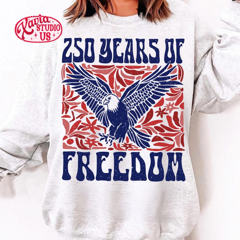 Puede incluir: Una sudadera blanca con un dise&ntilde;o patri&oacute;tico. El dise&ntilde;o incluye un &aacute;guila azul y blanca con las alas extendidas, rodeada de elementos florales rojos. El texto "250 YEARS OF FREEDOM" est&aacute; impreso en azul.