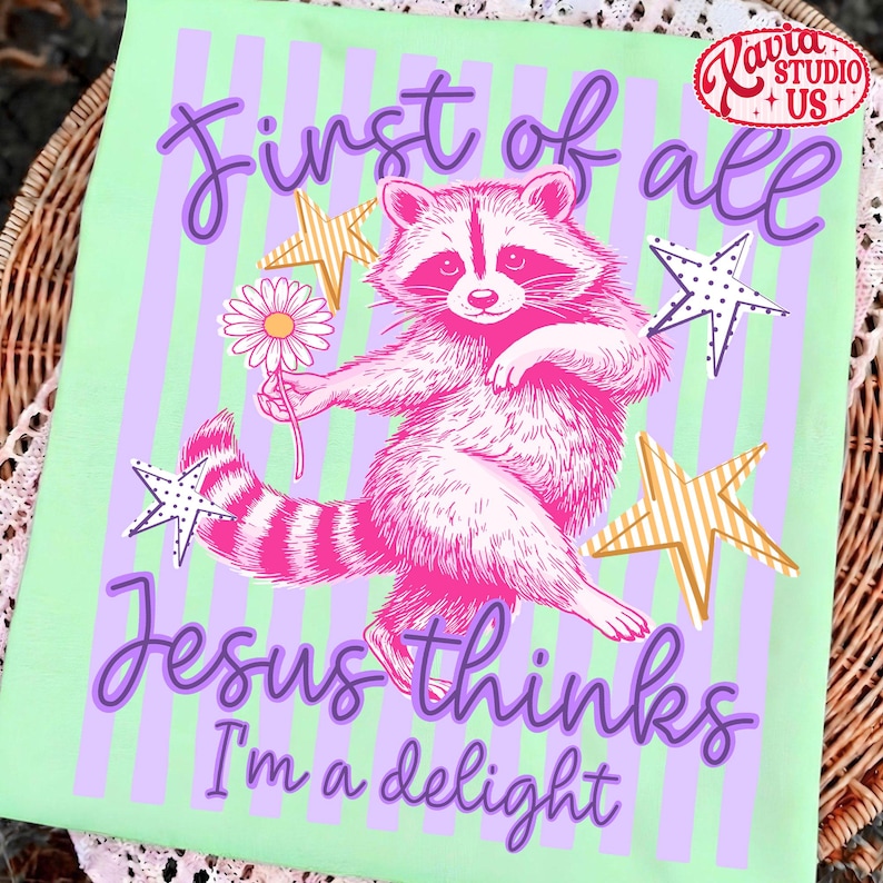 May include: A light green t-shirt with a pink raccoon holding a daisy, surrounded by stars. The text "First of all Jesus thinks I'm a delight" is written in purple script. A "Xavia Studio + US" logo is in the top right corner.
