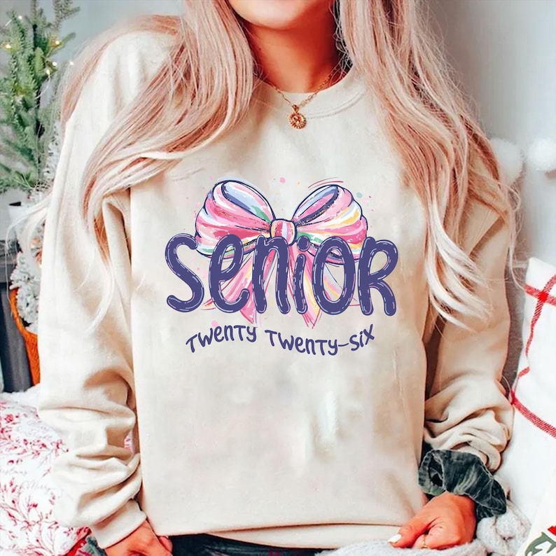 May include: A beige sweatshirt with the word "Senior" in blue script, above "Twenty Twenty-Six" in a smaller font. A colorful bow graphic is above the word "Senior". The sweatshirt is a neutral color.