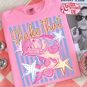 May include: Pink t-shirt with a graphic of a fish and the words "Shake That" in orange script. The design includes blue and white striped background and star accents. The shirt has a crew neck.