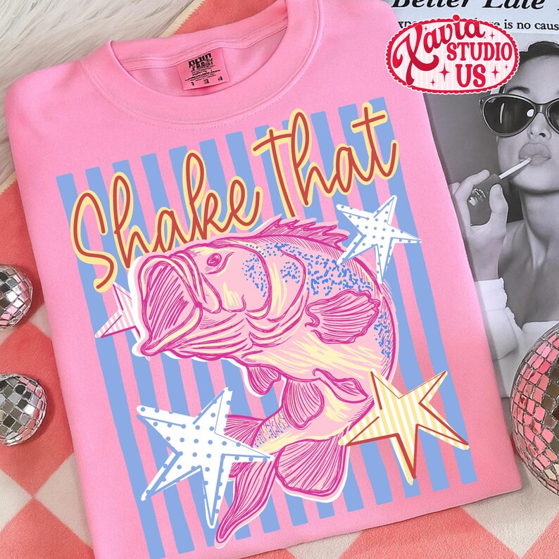 May include: Pink t-shirt with a graphic of a fish and the words "Shake That" in orange script. The design includes blue and white striped background and star accents. The shirt has a crew neck.