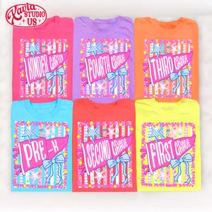 May include: Six short-sleeved t-shirts in pink, purple, orange, light blue, red, and yellow. Each shirt features a graphic of a pink pennant flag with a blue bow and stars, displaying grade levels: Kindergarten, Fourth, Third, Pre-K, Second, and First.