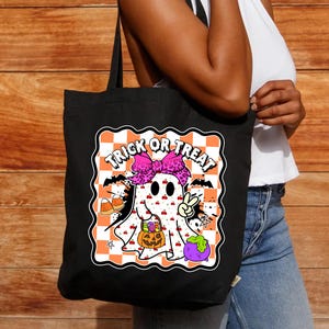 May include: Black tote bag featuring a Halloween-themed graphic. The design includes a cartoon ghost with a pink bow, holding a pumpkin and a peace sign, with the words "Trick or Treat" above. The background is a checkered pattern.