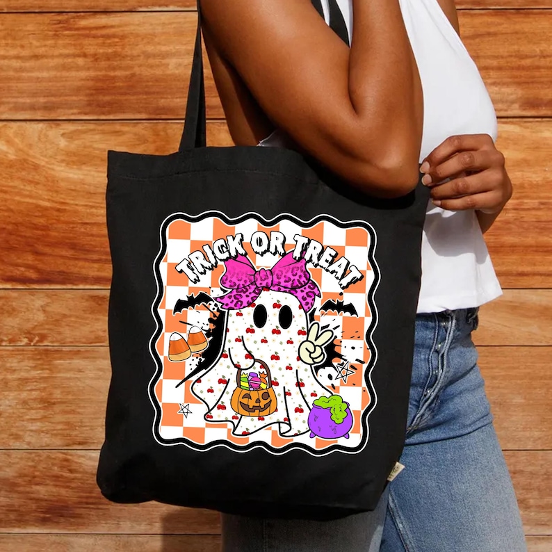 May include: Black tote bag featuring a Halloween-themed graphic. The design includes a cartoon ghost with a pink bow, holding a pumpkin and a peace sign, with the words "Trick or Treat" above. The background is a checkered pattern.
