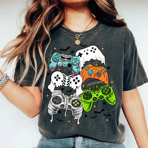 May include: Dark gray t-shirt featuring a colorful graphic of Halloween-themed video game controllers. The controllers are designed as a ghost, pumpkin, skeleton, and Frankenstein's monster. Bats and stars are also included in the design.