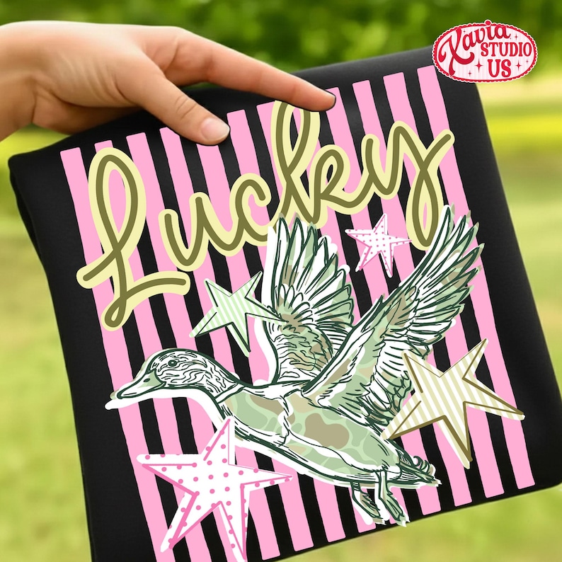 May include: Black t-shirt with pink vertical stripes and the word "Lucky" in a cursive font. A green and white illustration of a duck in flight is centered on the shirt, with star accents.