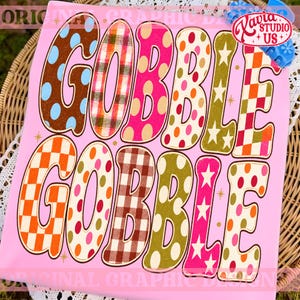 May include: A light pink t-shirt featuring the text GOBBLE GOBBLE in large, bold letters. Each letter displays a unique pattern, including polka dots, plaid, checkered prints, and stars in various colors like brown, orange, pink, and olive green.