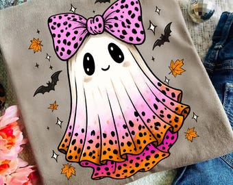 May include: A gray t-shirt featuring a cartoon ghost with a pink polka-dot bow. The ghost's cape transitions from white to pink and orange, with black polka dots. Bats, stars, and autumn leaves surround the ghost, creating a Halloween theme.
