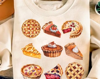 May include: Cream-colored sweatshirt featuring watercolor illustrations of various pies and pie slices. The designs include lattice-top pies, fruit pies, and pumpkin pie slices, showcasing a variety of fillings and toppings. The overall theme is centered around baked goods.