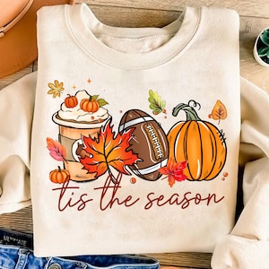 May include: Cream-colored sweatshirt with a fall-themed graphic. The design includes a coffee cup with whipped cream and pumpkins, a football, and a pumpkin. The text "tis the season" is written in a script font.