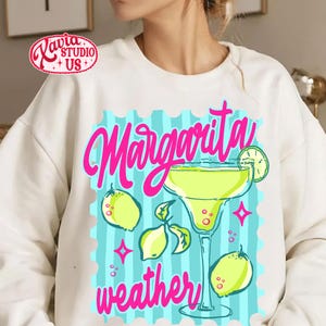 May include: Cream-coloured sweatshirt with a vibrant graphic. The design includes a margarita cocktail, lemons, and the words "Margarita weather" in pink script. The design is set against a blue background.