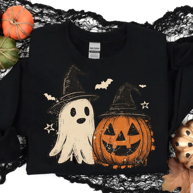 Op de afbeelding: Zwart sweatshirt met een Halloween-ontwerp. De afbeelding toont een spook met een heksenhoed naast een oranje pompoen met een heksenhoed. Vleermuizen en sterren zijn ook in het ontwerp opgenomen.