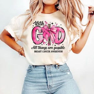 May include: Cream-colored t-shirt with a pink graphic design promoting breast cancer awareness. The design includes the words "With God" and "All things are possible" in black script, along with a pink ribbon and bow.