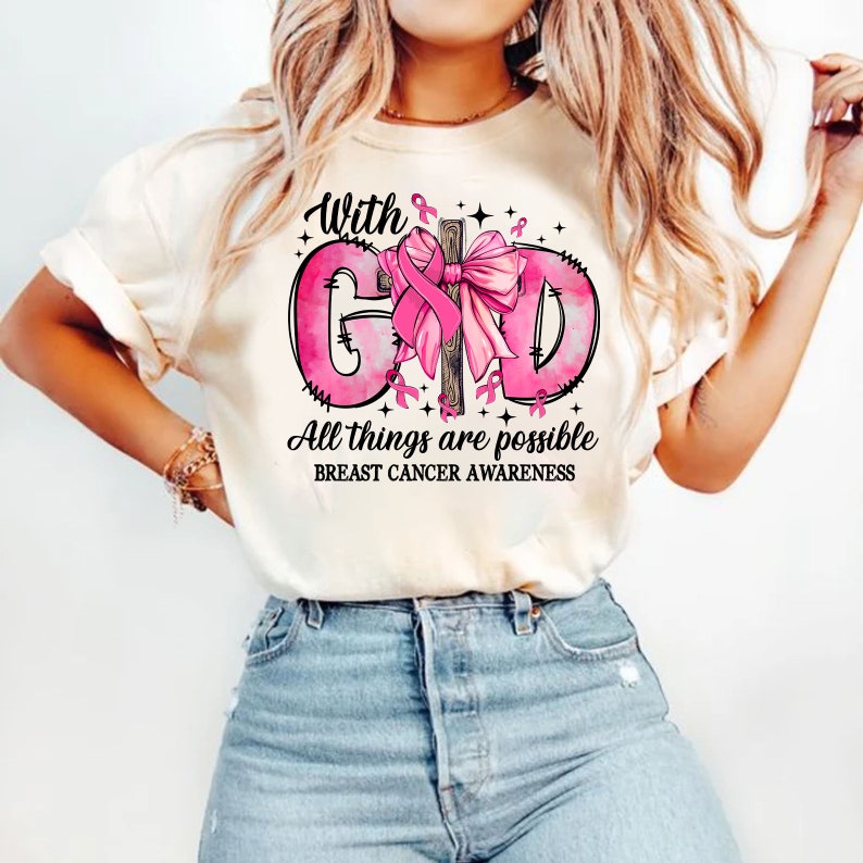 May include: Cream-colored t-shirt with a pink graphic design promoting breast cancer awareness. The design includes the words "With God" and "All things are possible" in black script, along with a pink ribbon and bow.