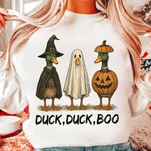 May include: White sweatshirt featuring three ducks dressed for Halloween. One duck wears a witch hat and cape, another is a ghost, and the third is a jack-o'-lantern. The text "DUCK, DUCK, BOO" is printed below the ducks.