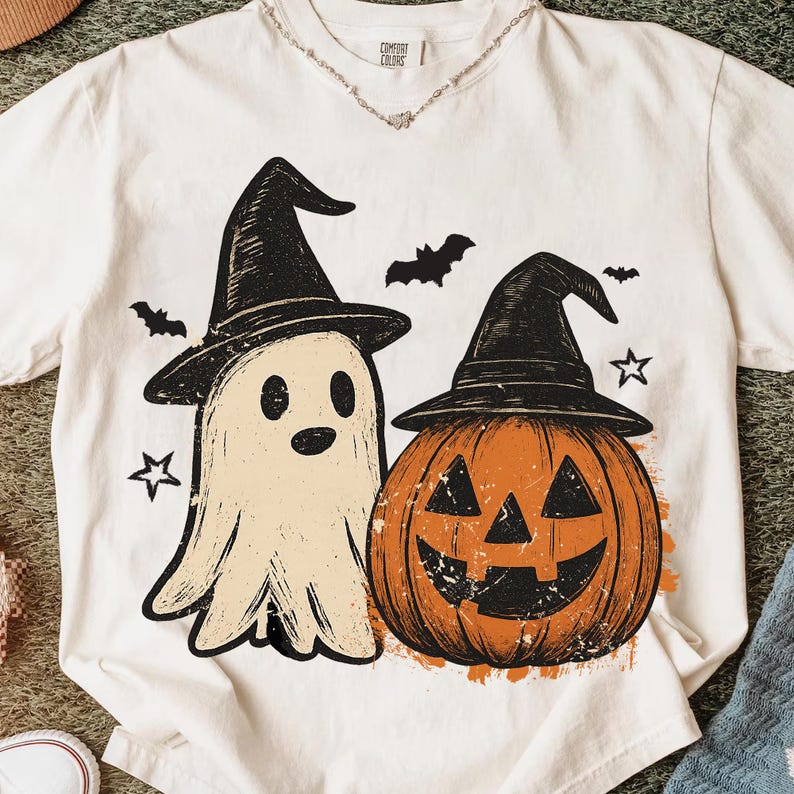 Op de afbeelding: Wit T-shirt met een Halloween-ontwerp. De afbeelding toont een spook met een heksenhoed, een pompoen met een heksenhoed, vleermuizen en sterren. Het shirt heeft het merklabel "Comfort Colors".