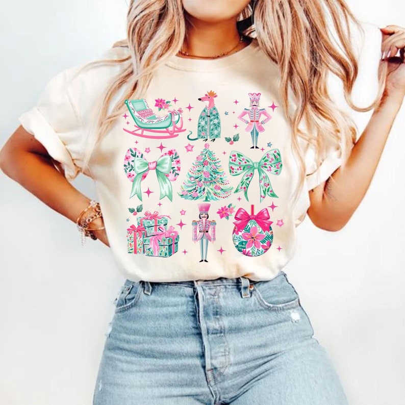 May include: Cream-colored t-shirt featuring a festive Christmas design. The design includes a sleigh, a reindeer, a nutcracker, bows, a Christmas tree, gift boxes, and a Christmas ornament. The illustrations are in shades of pink, green, and teal.