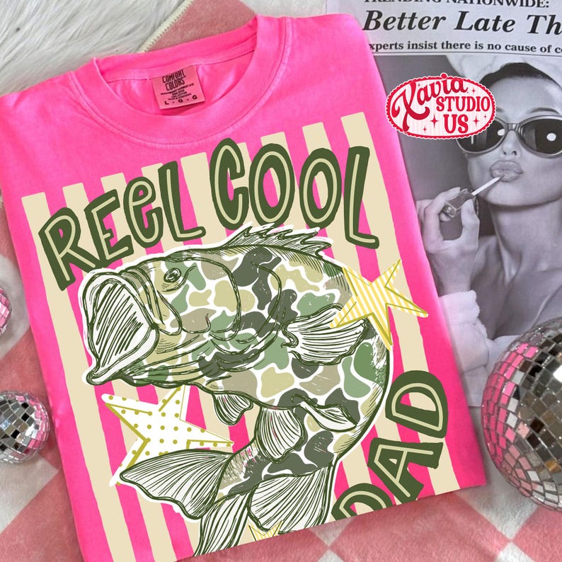 May include: A bright pink t-shirt with the words "REEL COOL DAD" in green and beige. The shirt features a fish with a camouflage pattern and two yellow star accents. The shirt is made of Comfort Colors material.