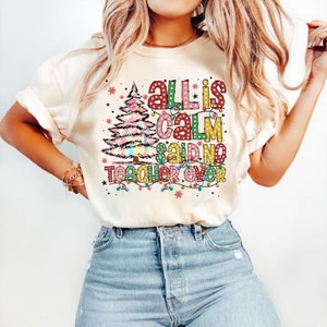 May include: Cream-colored t-shirt with the text "All is Calm Said No Teacher Ever" in colorful, polka-dotted letters and a Christmas tree graphic. The design includes string lights and snowflakes, creating a festive holiday theme.