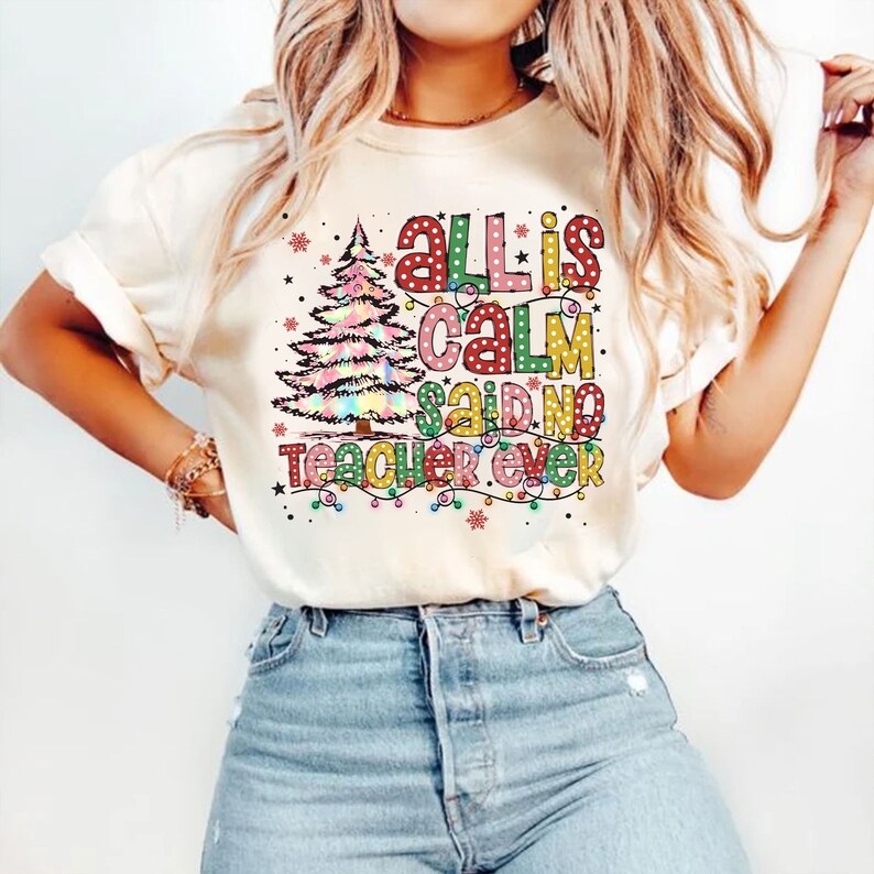 May include: Cream-colored t-shirt with the text "All is Calm Said No Teacher Ever" in colorful, polka-dotted letters and a Christmas tree graphic. The design includes string lights and snowflakes, creating a festive holiday theme.