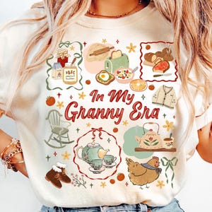 以下が含まれることがあります： クリーム色のTシャツにカラフルなグラフィックデザイン。デザインには、食べ物、ロッキングチェア、ティーポットのイラストと、赤いスクリプトで「In My Granny Era」というテキストが含まれています。全体的なテーマは、居心地の良いヴィンテージ美学です。
