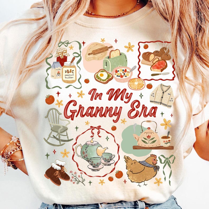 以下が含まれることがあります： クリーム色のTシャツにカラフルなグラフィックデザイン。デザインには、食べ物、ロッキングチェア、ティーポットのイラストと、赤いスクリプトで「In My Granny Era」というテキストが含まれています。全体的なテーマは、居心地の良いヴィンテージ美学です。