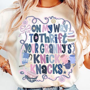 Peut inclure: T-shirt cr&egrave;me avec un motif color&eacute; indiquant "ON MY WAY TO THRIFT YOUR GRANNY'S KNICK KNACKS". Le design comprend des illustrations de tasses, de bols et d'autres objets vintage, dans un style fantaisiste et dessin&eacute; &agrave; la main.