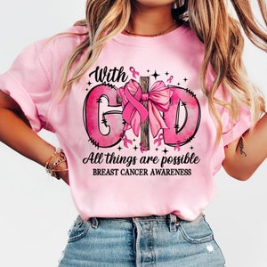 May include: A light pink t-shirt with the words "With God All things are possible" and "Breast Cancer Awareness" in black text. The design includes a pink ribbon and a cross with a pink bow. The shirt is a crew neck.