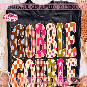 May include: A black t-shirt featuring the text GOBBLE GOBBLE in large, colorful, patterned letters. Each letter displays a unique design, including polka dots, plaid, checkered patterns, and stars in shades of pink, orange, brown, and green.