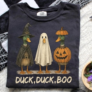 May include: Dark gray t-shirt featuring three ducks dressed for Halloween. One duck is a witch, one a ghost, and one a jack-o'-lantern. The text "DUCK, DUCK, BOO" is printed at the bottom in white.
