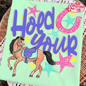 May include: A light green t-shirt with the words "Hold Your" in purple and a pink horseshoe. A cartoon horse with a blue saddle is featured below the text, surrounded by stars in pink, blue, and yellow.