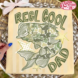 May include: Tan t-shirt with the text "REEL COOL DAD" in green. A fish with a camouflage pattern is in the center, with green and yellow star accents. The shirt has vertical stripes.