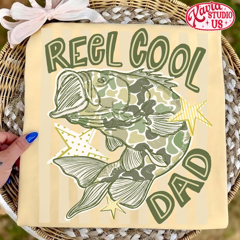 May include: Tan t-shirt with the text "REEL COOL DAD" in green. A fish with a camouflage pattern is in the center, with green and yellow star accents. The shirt has vertical stripes.