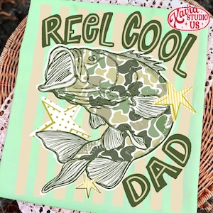 May include: A light green graphic featuring a fish with camouflage pattern, the words "REEL COOL DAD", and star accents. The design is on a light green background with vertical stripes.