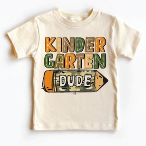 May include: A beige short-sleeved t-shirt with the text "KINDERGARTEN DUDE" in colorful letters. A cartoon pencil with a camouflage pattern is below the text.