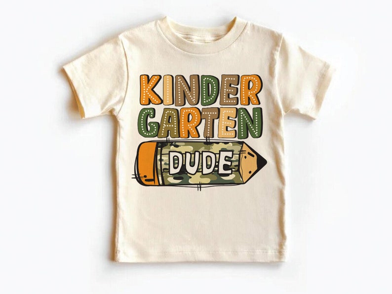 May include: A beige short-sleeved t-shirt with the text "KINDERGARTEN DUDE" in colorful letters. A cartoon pencil with a camouflage pattern is below the text.