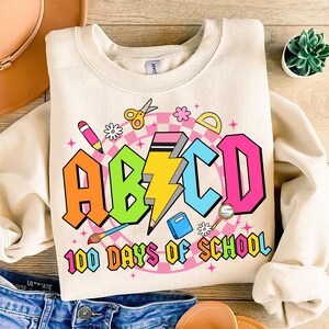 May include: A beige sweatshirt with a colorful graphic design featuring the letters "ABCD" and the text "100 Days of School". The graphic includes school supplies like a pencil, scissors, ruler, and paintbrush.