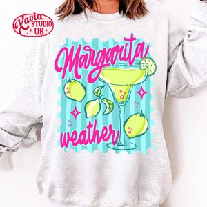 May include: A light grey sweatshirt with a vibrant graphic. The design includes the words "Margarita weather" in pink script, a margarita glass, lemons, and a lime slice. The background is light blue with vertical stripes.