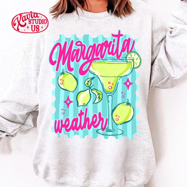 May include: A light grey sweatshirt with a vibrant graphic. The design includes the words "Margarita weather" in pink script, a margarita glass, lemons, and a lime slice. The background is light blue with vertical stripes.