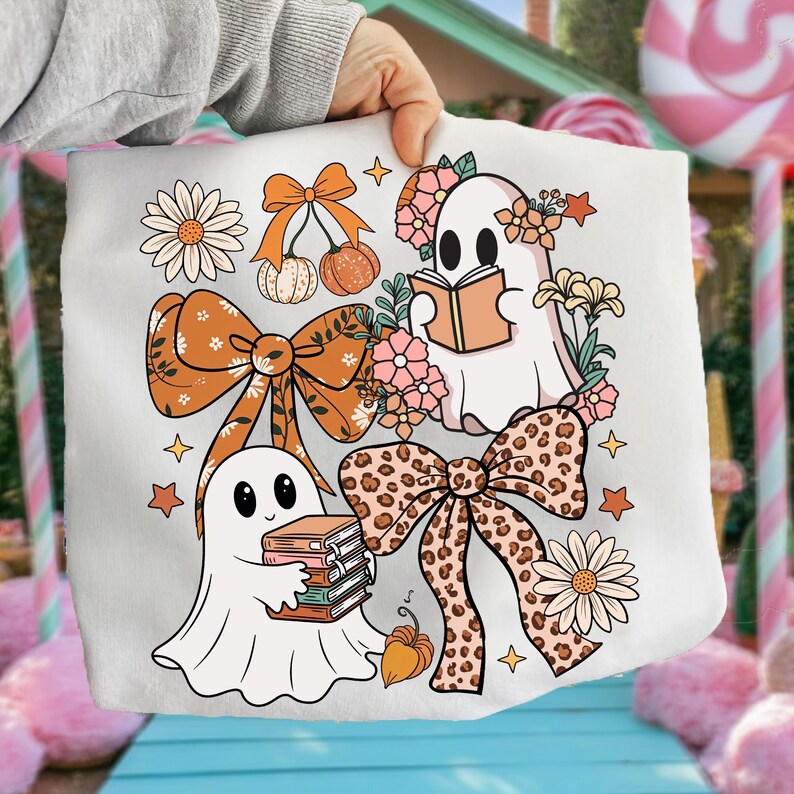 May include: White tote bag featuring a whimsical design with cartoon ghosts, bows, flowers, and pumpkins in shades of orange, peach, and brown. The ghosts are reading books. The design includes stars and daisy flowers.