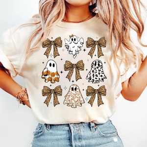 May include: Cream-colored t-shirt featuring a Halloween-themed design. The design includes nine ghost figures with various patterns, including leopard print and pumpkin faces, and leopard print bows. Small black stars are scattered around the ghosts.