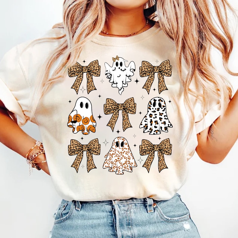 May include: Cream-colored t-shirt featuring a Halloween-themed design. The design includes nine ghost figures with various patterns, including leopard print and pumpkin faces, and leopard print bows. Small black stars are scattered around the ghosts.