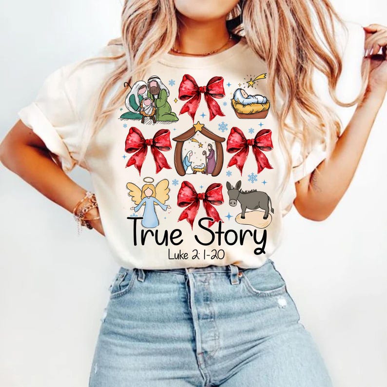 May include: Cream-colored t-shirt featuring a colorful graphic of a nativity scene, angels, and red bows. The text "True Story" and "Luke 2:1-20" are printed below the illustrations. The shirt is worn by a person.